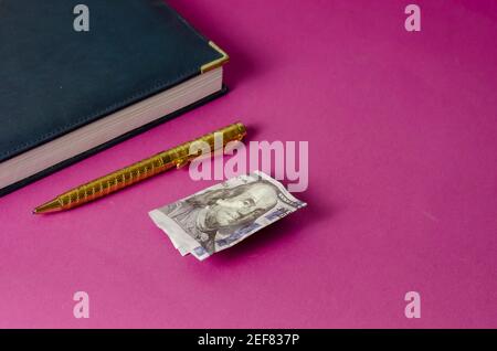 Journal, stylo or et cent dollars américains sur fond rose. Vue de dessus inclinée. Mise au point sélective Banque D'Images