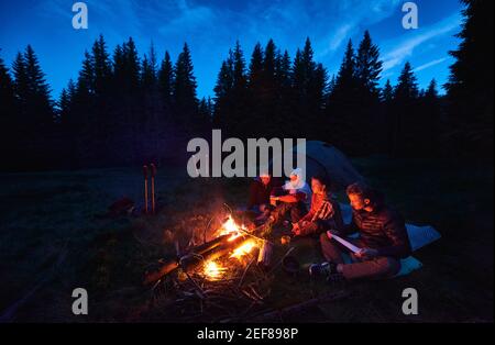 Groupe de quatre touristes assis près du feu de camp, en profitant de l'air frais près de la tente avec des équipements de touristes à proximité, sombre atmosphère relaxante. Camping en soirée près du feu, forêt d'épicéa en arrière-plan. Banque D'Images