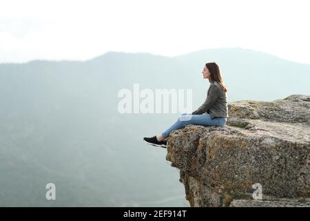 Profil complet d'une femme pensive assise contemplant des vues au sommet d'une falaise dans la montagne Banque D'Images