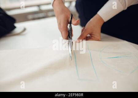 Gros plan. Mains femme tailleur travaillant la découpe d'un rouleau de tissu sur lequel elle a marqué le modèle du vêtement qu'elle fabrique avec la craie de tailleurs. Banque D'Images