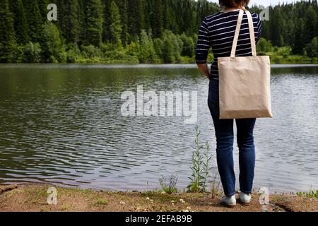 Femme en t-shirt rayé portant vide réutilisable sac maquette, vue sur la rivière. Maquette de sac fourre-tout en toile écologique pour femme Banque D'Images