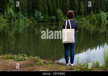 Femme debout sur la rive du lac portant une maquette de sac à provisions réutilisable vide. Maquette de sac fourre-tout en toile écologique pour femme Banque D'Images