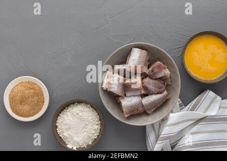 Dans un bol, des morceaux de merlu et des produits pour la cuisine frite, des œufs, de la farine et de la chapelure ont été présentés avec une serviette de cuisine sur fond gris Banque D'Images