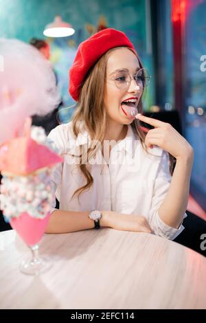 Une française en béret rouge mange un dessert sucré de bonbons en coton rose, un gâteau et un milk-shake avec son doigt. Banque D'Images