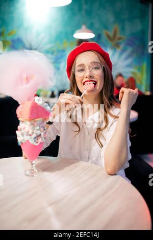 Jeune femme en béret rouge et verres a un grand dessert sucré avec des bonbons en coton rose dans un café en France. Banque D'Images