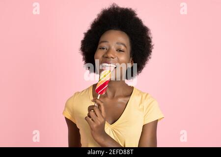 Portrait de la jeune femme positive à la peau sombre avec des cheveux afro, léchant délicieux Lollipop coloré, portant un t-shirt jaune d'été, posant sur le rose Banque D'Images