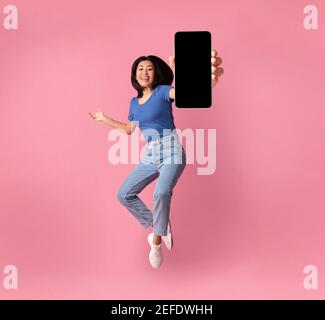 Bonne jeune femme asiatique montrant un écran de smartphone vide tout en sautant sur fond rose studio, collage, photo pleine taille. Je le recommande vivement Banque D'Images