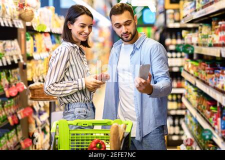 Portrait de la jeune famille faire des courses ensemble dans un supermarché. Couple souriant utilisant le téléphone balayer les produits alimentaires dans les allées du magasin Ind Banque D'Images