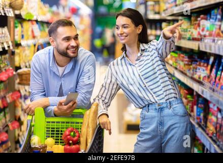 Je veux ça. Un couple millénaire faisant de l'épicerie magasiner ensemble, un homme se penchant sur un chariot et utilisant un téléphone portable, une femme pointant sur une étagère, une famille choisissant F Banque D'Images