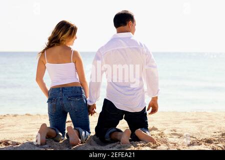 Vue arrière d'un homme adulte moyen et d'un jeune femme à genoux en tenant les mains sur la plage Banque D'Images