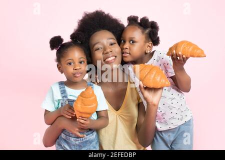 Gros plan portrait de famille de jeune femme africaine attrayante maman et de deux petites filles mignonnes, s'amusant ensemble, manger des croissants frais Banque D'Images