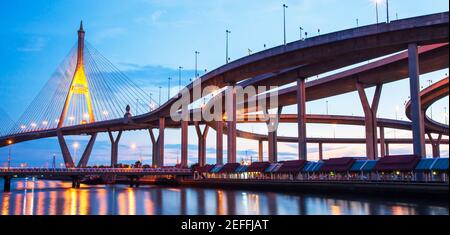 Vue pittoresque sous-jacente de l'échangeur d'autoroute et des ponts suspendus au crépuscule, reflet lumineux sur une rivière. Pont Bhumibol, Thaïlande. Banque D'Images