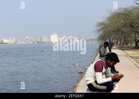 Un jeune garçon indien regarde son téléphone portable tout en s'asseyant près du lac Horseshoe Bhalswa à Delhi, en Inde, pendant un après-midi d'hiver ensoleillé Banque D'Images