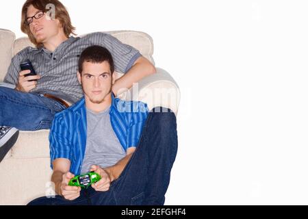 Portrait of a young man playing video game avec une télécommande et un autre jeune homme à l'aide d'un téléphone mobile derrière lui Banque D'Images