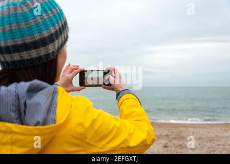 femme portant un manteau jaune prenant une photo de la mer avec son smartphone. Scène Moody. Banque D'Images