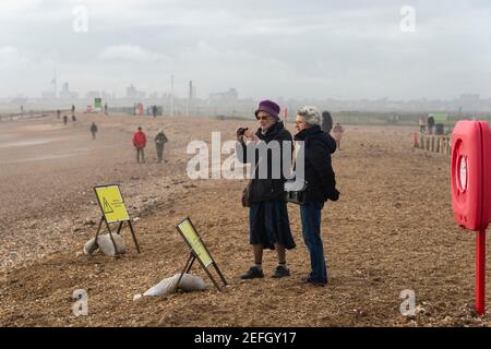 Une femme âgée et son amie prennent une photo avec un appareil photo numérique compact, debout sur la plage de galets en hiver. Banque D'Images