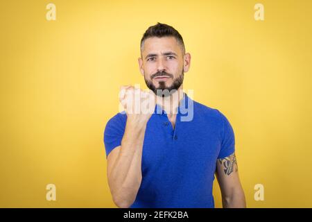 Beau homme avec barbe portant un polo bleu sur fond jaune en colère et fou levant poing frustré et furieux tout en criant avec colère. RA Banque D'Images