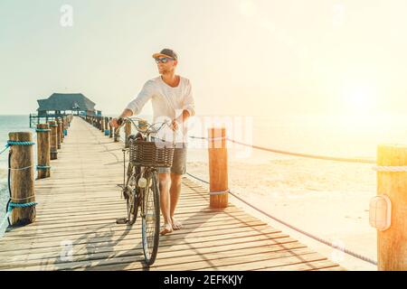 Portrait d'un homme heureux souriant pieds nus vêtu de vêtements d'été légers et de lunettes de soleil à vélo sur la jetée en bois. Des vacances imprudentes Banque D'Images