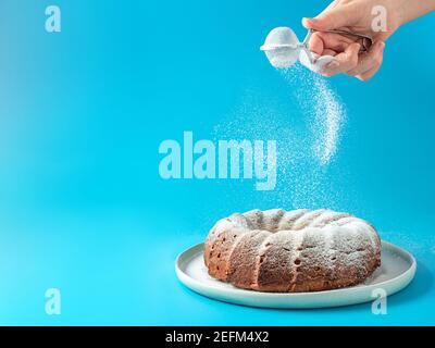 La main de la femme saupoudrant de sucre glace sur la maison fraîche fait du bundt gâteau. Le sucre en poudre tombe sur un gâteau de pain frais parfait sur fond bleu. Espace de copie pour le texte. Idées et recettes pour le petit déjeuner ou le dessert Banque D'Images