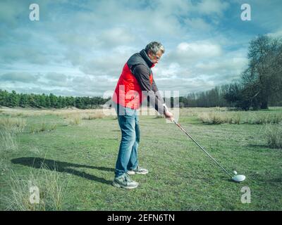 Senior citoyen pratiquant son swing de golf sur un terrain rustique par temps nuageux Banque D'Images