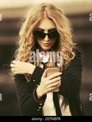Femme d'affaires blonde utilisant un téléphone cellulaire dans la rue de ville élégante modèle de mode avec poils longs et bouclés en veste noire et lunettes de soleil Banque D'Images