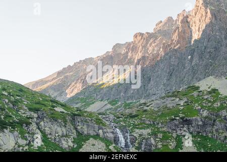 Réflexions dans l'eau calme du lac, cascade et montagnes au coucher du soleil. Banque D'Images
