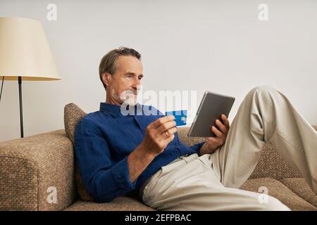 Beau barbu homme tenant une tablette numérique et une carte de crédit assis sur un canapé Banque D'Images
