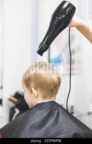 Salon de coiffure dans salon de beauté sèche cheveux avec grand sèche cheveux noir après avoir coupé la coupe de cheveux pour enfant, garçon, caucasien, blond. Tir vertical. Banque D'Images