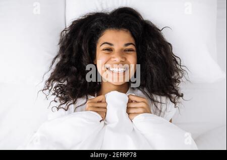 Matin du week-end. Vue de dessus portrait d'une belle fille se relaxant dans un lit et souriant. Une femme afro-américaine satisfaite se réveillant, se sentant fraîche et gaie le matin, le week-end à la maison Banque D'Images