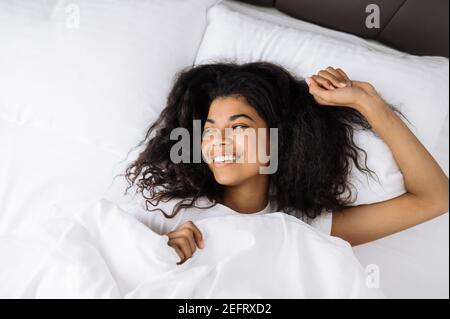 Souriante multiraciale noire curly fille utilise le téléphone cellulaire, parler avec des amis ou la famille pendant que s'assoit sur le canapé dans le salon et regarde la caméra, le concept de communication Banque D'Images