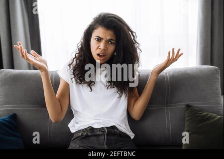 Souriante multiraciale noire curly fille utilise le téléphone cellulaire, parler avec des amis ou la famille pendant que s'assoit sur le canapé dans le salon et regarde la caméra, le concept de communication Banque D'Images