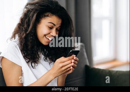 Adolescente afro-américaine jolie fille s'assoit au canapé dans le salon et utilise son smartphone. Belle fille en tenue décontractée passe du temps à la maison, à discuter avec des amis ou la famille, à surfer sur Internet Banque D'Images