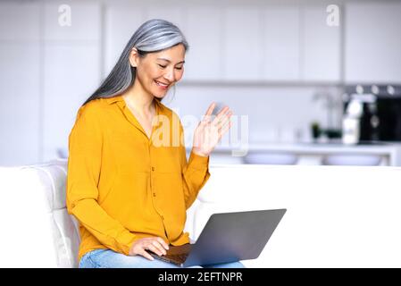 Une femme asiatique souriante et chaleureuse, assise sur le canapé dans le salon, utilisant un ordinateur portable, tout en travaillant à distance, communique avec des collègues ou des amis d'affaires, fait des vagues, salue Banque D'Images