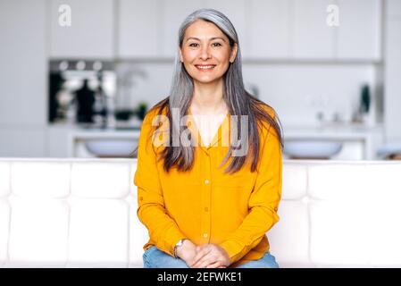 Portrait d'une charmante femme asiatique senior à cheveux longs gris vêtue de vêtements élégants et décontractés, assise sur un canapé dans le salon, regardant directement l'appareil photo avec un sourire amical Banque D'Images