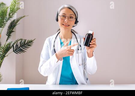 Un médecin asiatique amical aux cheveux gris, vêté d'un uniforme médical et d'un casque, présente un médicament contre le rhume à un patient lors d'une consultation en ligne. Assistance médicale à distance Banque D'Images