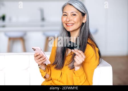 Bonne femme d'âge moyen souriante aux cheveux gris, fait du shopping en ligne. Joyeuse femme asiatique senior tient la carte de crédit et son smartphone, shopping en ligne, paiement en ligne, assis dans le salon Banque D'Images