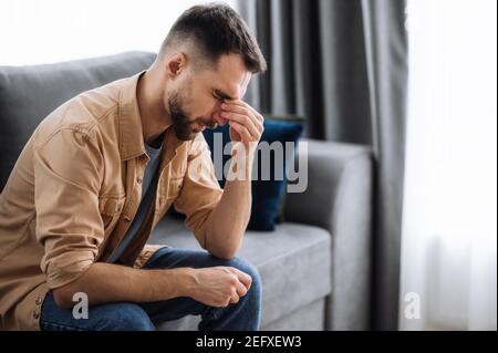 Un gars fatigué assis sur le canapé à la maison, ayant un mal de tête terrible. Exténué massage masculin caucasien les yeux, se sentir malsain, attraper un rhume ou avoir la grippe, bouleversé au sujet de la maladie Banque D'Images