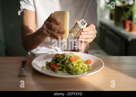 Gros plan des mains de femme râpant le parmesan dans les pâtes avec sauce pesto, tomates cerises fraîches. Banque D'Images