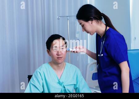 Jeune femme médecin avec stéthoscope examinant les oreilles de l'Asie patient mâle en uniforme utilisant un thermomètre tympanique pour la température dans hôpital Banque D'Images