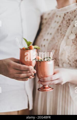 Des mains de jeunes couples amoureux se trinquer des gobelets en laiton avec un cocktail de glace avec des agrumes et des pailles en verre écologiques ensemble, foyer sélectif. Anniversaire de mariage, concept de la Saint-Valentin Banque D'Images