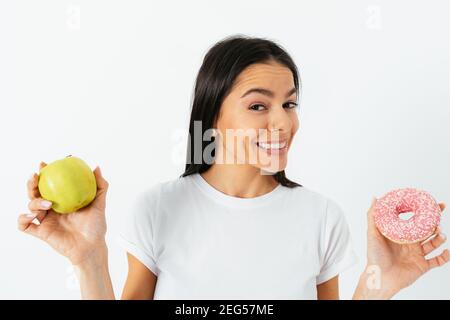 Gros plan portrait jeune femme tenant le donut et la pomme dans ses mains smily smily et regardant l'appareil photo, sain contre le concept de nourriture indésirable. Banque D'Images