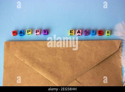 Happy Easter lettrage texte composé de lettres en bois multicolores, enveloppe marron et plume blanche sur un fond bleu clair. Des vacances minimalistes de retour Banque D'Images