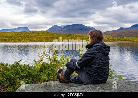 Femme assise sur un rocher au bord d'un lac en automne avec Papporten en arrière-plan, Abisko, Laponie suédoise, Suède Banque D'Images