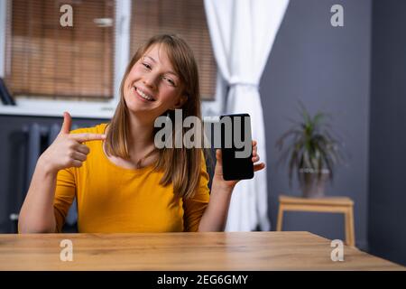 Femme blonde au bureau à domicile, souriante, tenant un smartphone moderne avec un écran maquette vierge, la femme adolescente pointe avec son index vers elle. Vide Banque D'Images