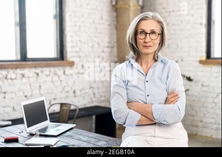 Femme d'affaires âgée sérieuse et stricte portant une chemise élégante et décontractée et des supports de lunettes élégants avec les bras croisés dans un espace de bureau moderne. A Banque D'Images