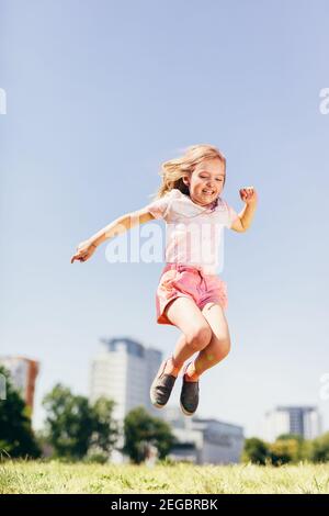 Petite fille heureuse sautant haut sur la prairie. L'été dans la ville. Enfance. Banque D'Images