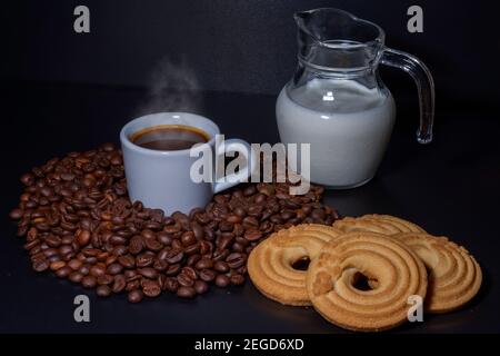 Tasse de café blanc avec une boisson à l'intérieur et entourée de grains de café torréfiés de la variété Robusta, un pot en verre avec du lait à l'intérieur et un beignet de CA Banque D'Images