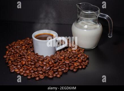 Tasse de café blanc avec une boisson à l'intérieur et entourée de grains de café torréfiés de la variété Robusta et d'une verseuse en verre avec lait à l'intérieur. Petit déjeuner Banque D'Images