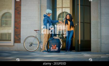 Happy Food Delivery un homme portant un sac à dos isotherme fait du vélo pour livrer une commande de restaurant à une belle cliente féminine. Le service de messagerie livre le déjeuner à Banque D'Images