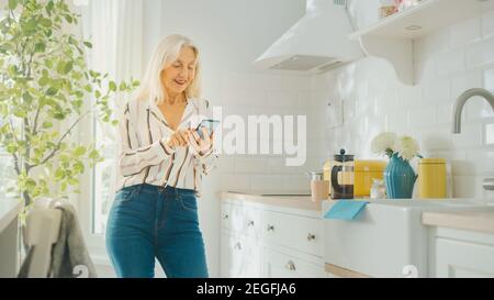 Authentique femme senior en Jean bleu et blouse à rayures blanches utilise un smartphone dans la cuisine. Bonne belle retraite détente à la maison. Banque D'Images